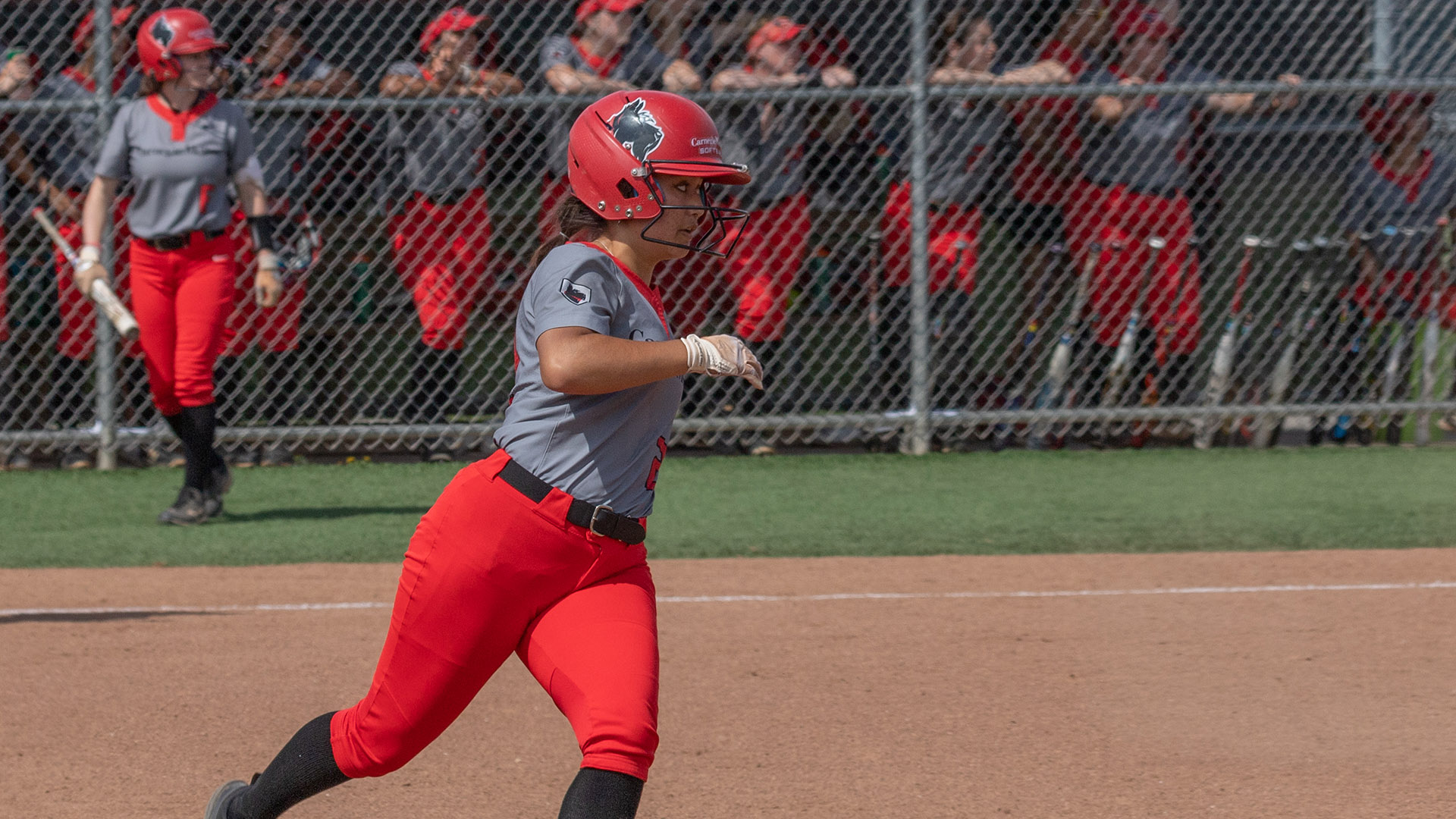 women's softball player wearing gray top and red pants looks to round first base with teammates watching from the dugout