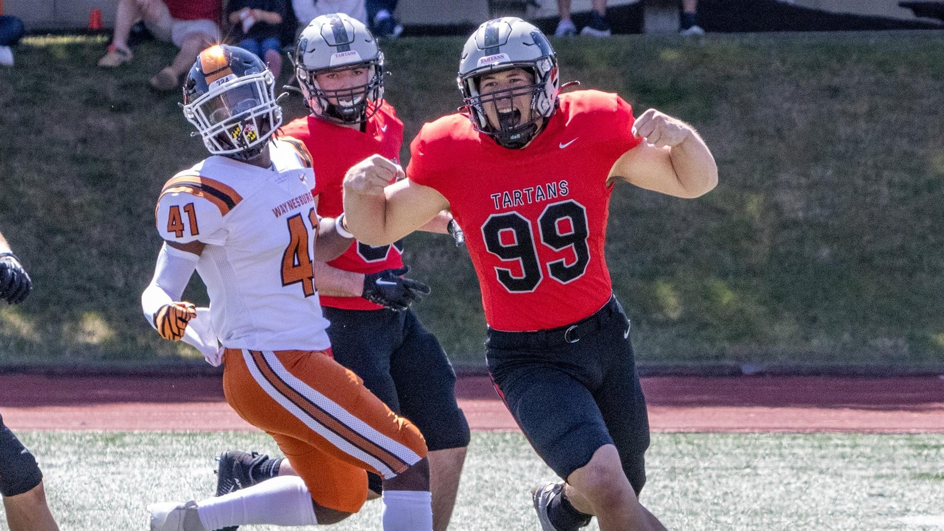 football player wearing a red jersey celebrates on the field with a teammate and opposing player in the background