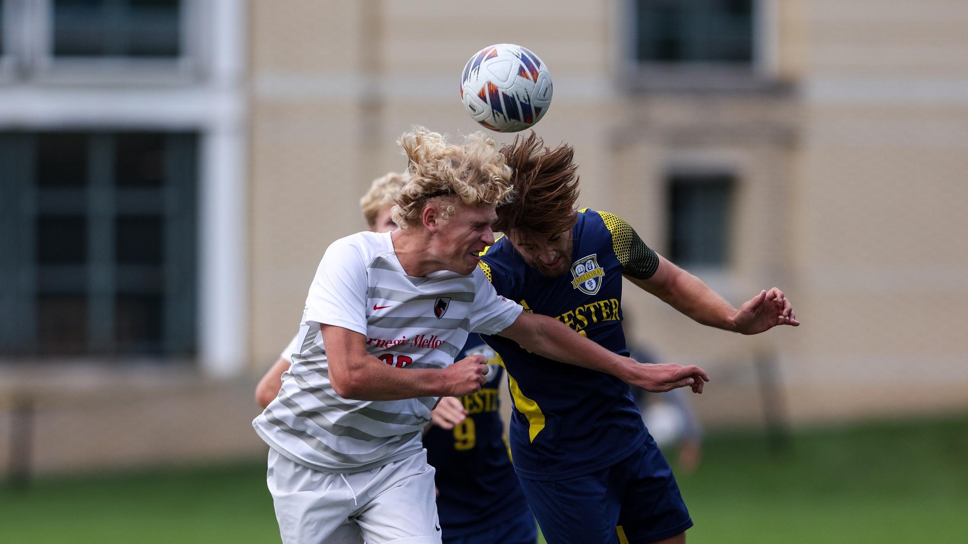 men's soccer player wearing a white uniform jumps to head the ball with a player wearing a blue uniform jumping with him