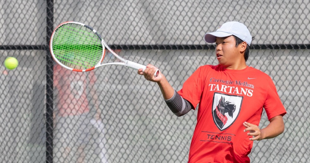 men's tennis player wearing a red shirt and white hat swings at a ball with his right hand holding the racket