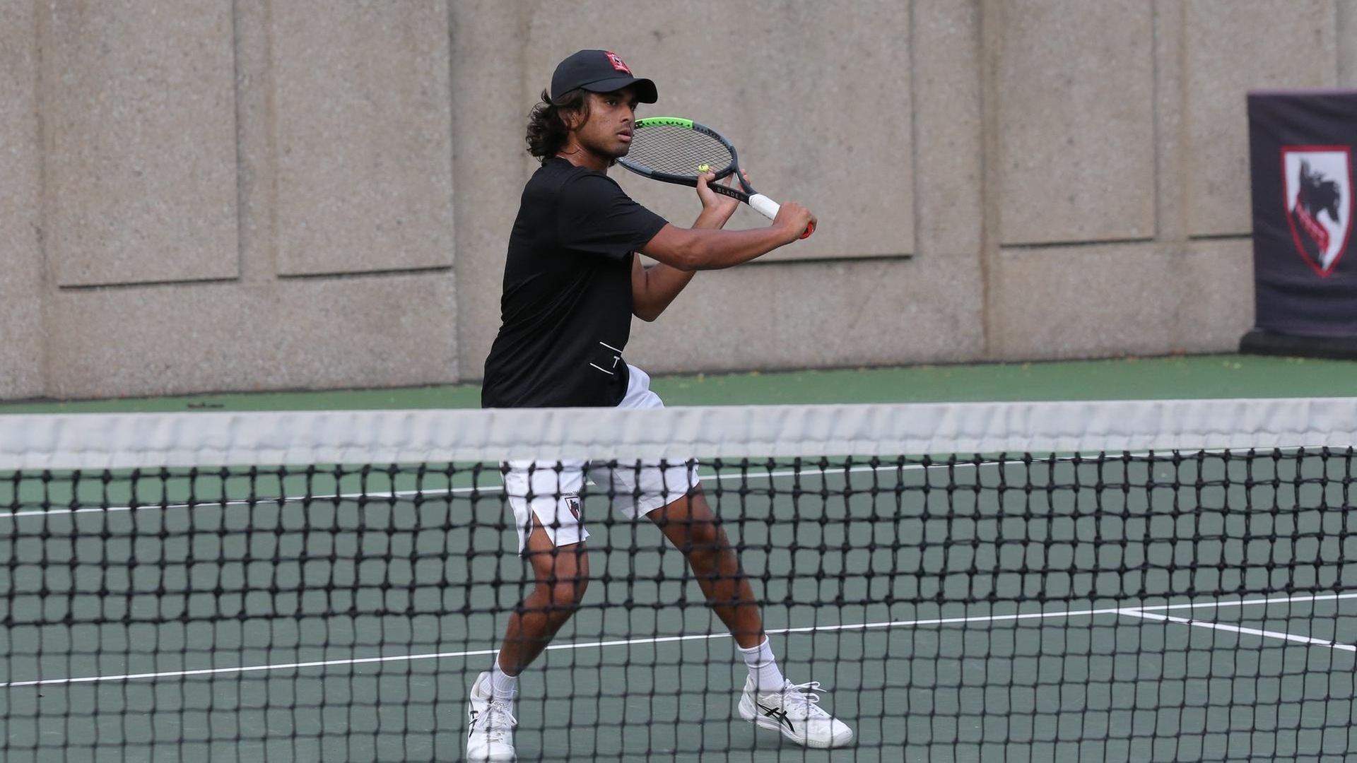 men's tennis player wearing a black hat, black shirt, and white shorts swings a racquet