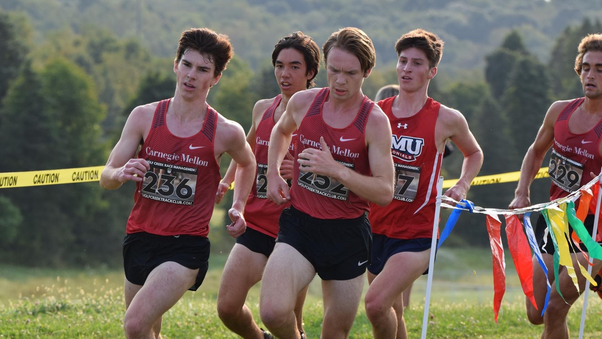 five men's cross country runners rounding a corner with trees blended in the background