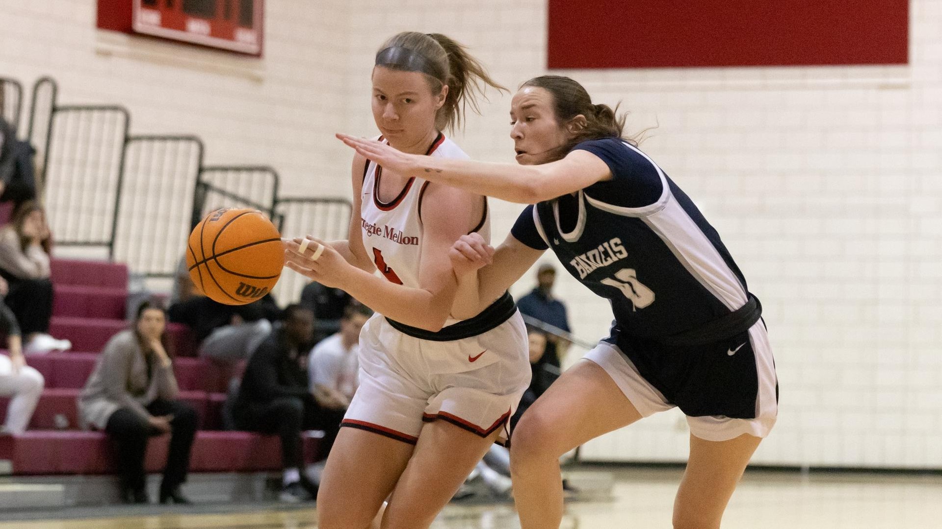 women's basketball player wearing a white uniform dribbles ball past a player wearing a blue uniform