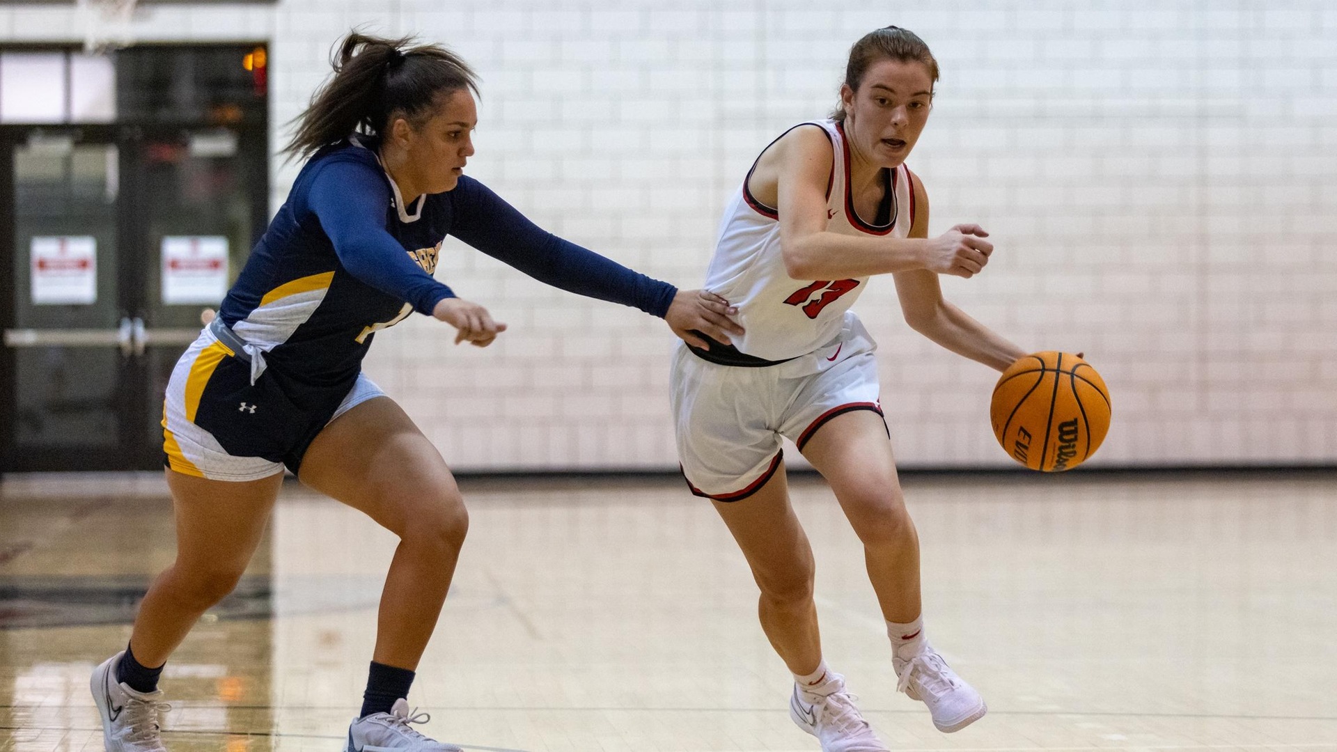 women's basketball player wearing a white uniform dribbles with her left hand by a player wearing a blue uniform