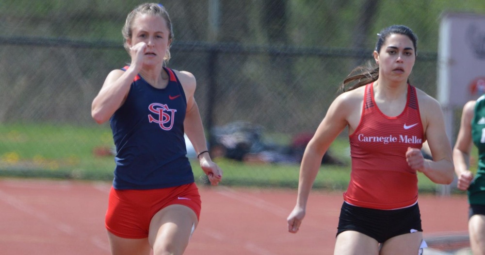 female track runners in a sprint, one wearing a blue top and red bottoms, one wearing a red top and black bottoms